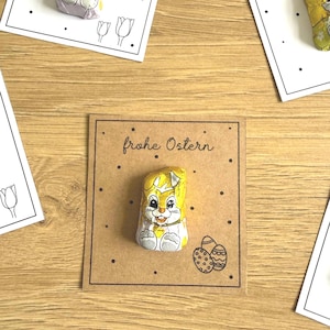 Può includere: Primo piano di coniglietti di cioccolato pasquali avvolti in carta stagnola gialla. I coniglietti sono posizionati su biglietti con la scritta "frohe Ostern" e illustrazioni di tulipani e uova di Pasqua. Sfondo in legno marrone chiaro.