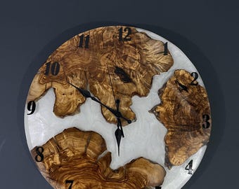 May include: A round wooden clock with white resin accents. The clock face has black Roman numeral markings and black hands. The wood grain is visible and has a natural brown color.