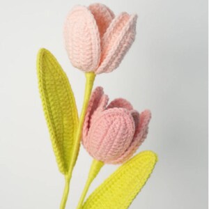 May include: Two handmade crochet tulips with pink petals and bright yellow-green leaves. The stems are yellow. The flowers are set against a plain white background.