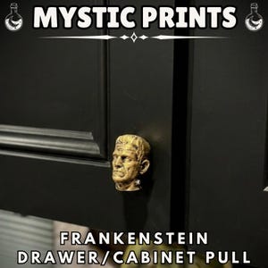 Pode incluir: Um puxador de gaveta/armário Frankenstein dourado está montado em uma porta de armário preta. O puxador é uma escultura detalhada da cabeça de Frankenstein. O texto "MYSTIC PRINTS" está no topo e "FRANKENSTEIN DRAWER/CABINET PULL" na parte inferior.