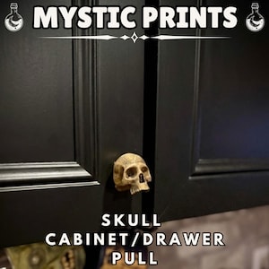 May include: A black cabinet door with a skull-shaped handle. The handle is a light tan colour and is mounted on the door. The text "MYSTIC PRINTS" is at the top, and "SKULL CABINET/DRAWER PULL" is at the bottom.