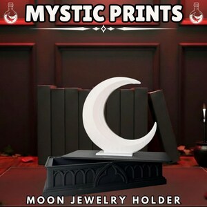 May include: A white crescent moon jewelry holder with the text "MOON JEWELRY HOLDER" below. The holder sits on a black rectangular base above a black coffin-shaped jewelry box. The words "MYSTIC PRINTS" are at the top.
