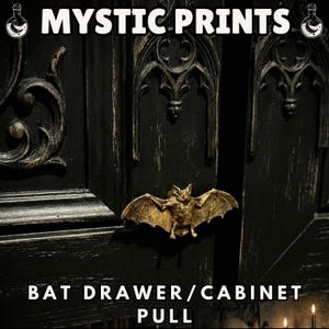 May include: A gold-coloured bat drawer pull is mounted on a black cabinet door. The cabinet door has ornate gothic-style carvings. The text "MYSTIC PRINTS" is at the top, and "BAT DRAWER/CABINET PULL" is at the bottom.