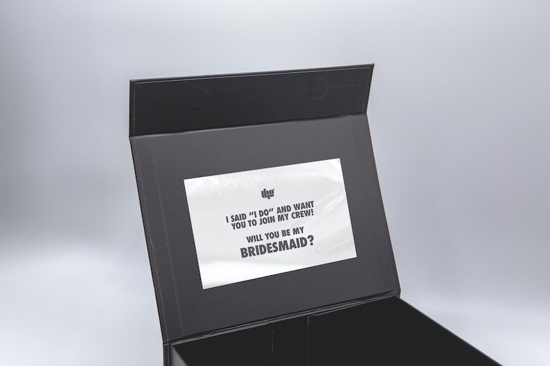 May include: A black box with a hinged lid, featuring a white rectangular label inside. The label has text that reads, "I SAID 'I DO' AND WANT YOU TO JOIN MY CREW! WILL YOU BE MY BRIDESMAID?" The box is open.