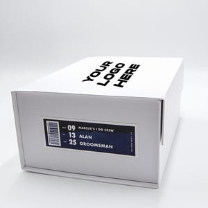 May include: A white box with the text "YOUR LOGO HERE" printed on the top. A label on the side reads "MARCUS'S I DO CREW", with the names "ALAN" and "GROOMSMAN" listed below. The box is on a white surface.