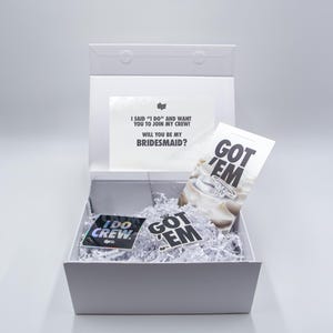 May include: A white gift box with a lid, filled with white paper shreds. Inside are cards and stickers with text: "I SAID 'I DO' AND WANT YOU TO JOIN MY CREW! WILL YOU BE MY BRIDESMAID?" and "GOT 'EM" and "I DO CREW."