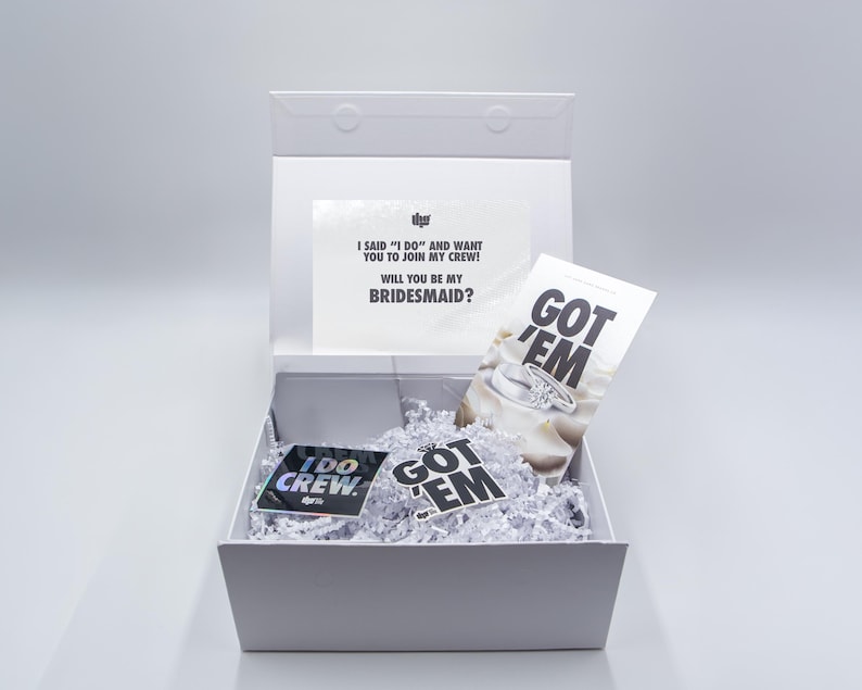 May include: A white gift box with a lid, filled with white paper shreds. Inside are cards and stickers with text: "I SAID 'I DO' AND WANT YOU TO JOIN MY CREW! WILL YOU BE MY BRIDESMAID?" and "GOT 'EM" and "I DO CREW."