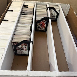 May include: A cardboard box with white dividers, filled with stacks of trading cards. Black card holders separate the stacks. Some cards feature hockey players in action. The box is open, revealing the contents.
