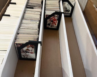 May include: A cardboard box with white dividers, filled with stacks of trading cards. Black card holders separate the stacks. Some cards feature hockey players in action. The box is open, revealing the contents.