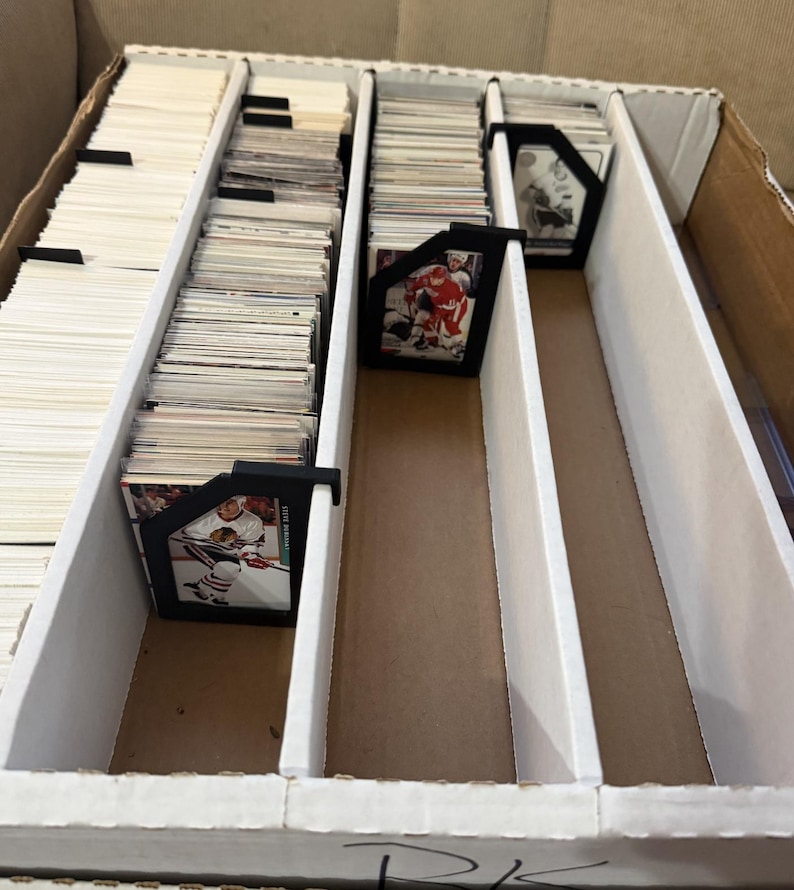 Puede incluir: Una caja de cart&oacute;n con divisores blancos, llena de montones de cromos. Los soportes negros para tarjetas separan las pilas. Algunas tarjetas presentan jugadores de hockey en acci&oacute;n. La caja est&aacute; abierta, revelando el contenido.
