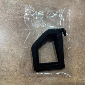 Puede incluir: Un componente de pl&aacute;stico negro, posiblemente un soporte o montaje, est&aacute; encerrado en una bolsa de pl&aacute;stico transparente. El componente tiene una forma geom&eacute;trica con una base rectangular y una parte superior en &aacute;ngulo, con una peque&ntilde;a pesta&ntilde;a saliente.