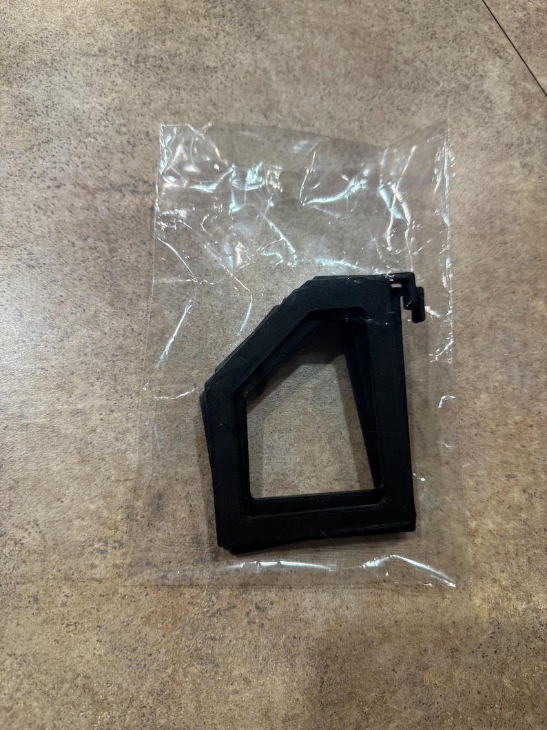 Puede incluir: Un componente de pl&aacute;stico negro, posiblemente un soporte o montaje, est&aacute; encerrado en una bolsa de pl&aacute;stico transparente. El componente tiene una forma geom&eacute;trica con una base rectangular y una parte superior en &aacute;ngulo, con una peque&ntilde;a pesta&ntilde;a saliente.