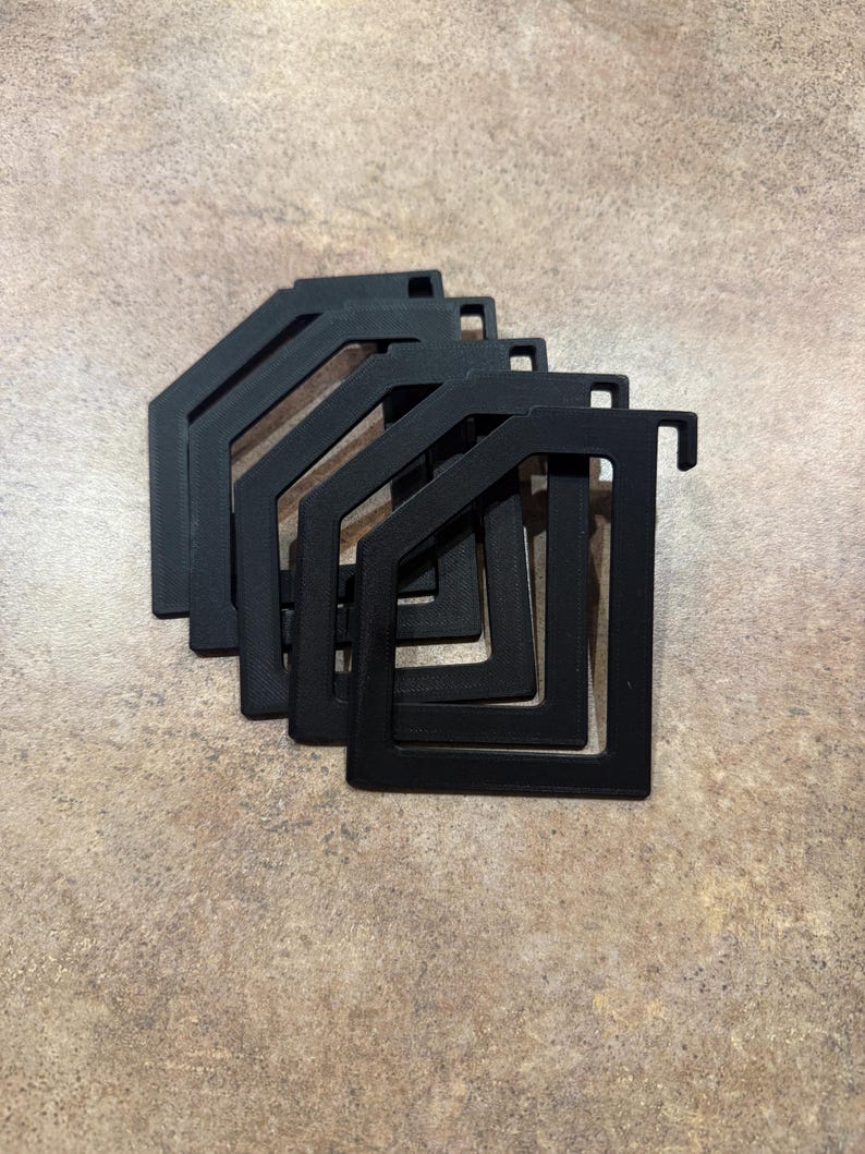 Puede incluir: Una pila de cinco objetos negros de forma geom&eacute;trica. Cada objeto tiene un marco rectangular con un gancho en un lado y un recorte rectangular m&aacute;s peque&ntilde;o. Los objetos est&aacute;n dispuestos en un patr&oacute;n ligeramente superpuesto sobre una superficie marr&oacute;n moteada.
