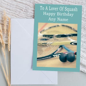 Puede incluir: Una tarjeta de cumpleaños turquesa con el texto "To A Lover Of Squash Happy Birthday Any Name" se muestra con un sobre blanco. La tarjeta presenta raquetas y pelotas de squash. El texto "Personalise With A Name" está debajo.