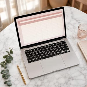 May include: A silver laptop with a black keyboard and trackpad displays a digital spreadsheet template featuring a soft pink header bar and grid lines on its screen. The device is positioned on a white marble surface with a gold pen and green leaves.