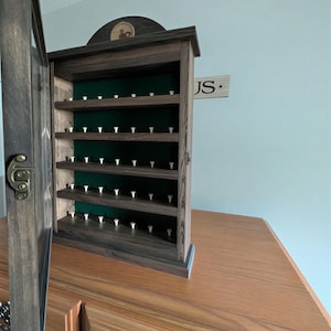 May include: A dark stained wooden display cabinet with multiple shelves, holding numerous white golf tees. The cabinet has a green felt backing and a hinged door with a metal latch. A small wooden sign with the initials "JS" is visible.