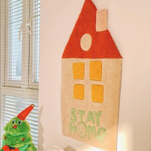 May include: A beige and orange felt house-shaped wall hanging with the words "STAY HOME" in green felt.