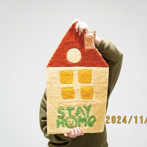 May include: A person holds a yellow and orange felt house with a red roof. The house has four yellow windows and the words "STAY HOME" are embroidered in green on the front. The date "2024/11/17" is written in orange on the bottom right corner.