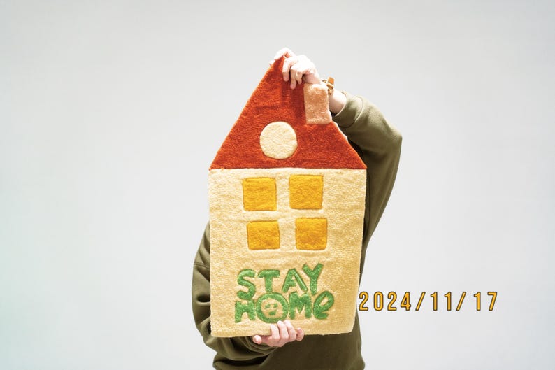 May include: A person holds a yellow and orange felt house with a red roof. The house has four yellow windows and the words "STAY HOME" are embroidered in green on the front. The date "2024/11/17" is written in orange on the bottom right corner.