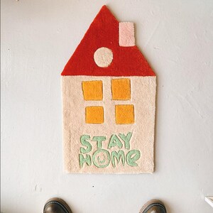 May include: A beige and red house-shaped rug with yellow windows and the words "STAY HOME" in green. The rug has a pink chimney and a white circle on the roof.