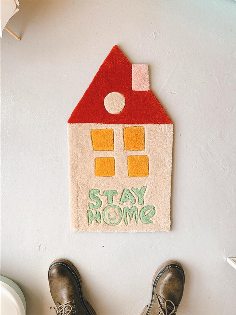 May include: A beige and red house-shaped rug with yellow windows and the words "STAY HOME" in green. The rug has a pink chimney and a white circle on the roof.