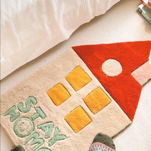 May include: A beige and cream rug shaped like a house with a red roof and yellow windows. The rug has the words "Stay Home" written in green on it.