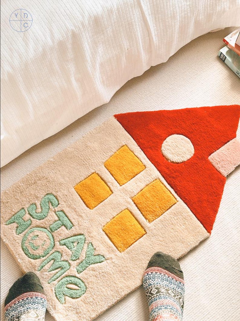 May include: A beige and cream rug shaped like a house with a red roof and yellow windows. The rug has the words "Stay Home" written in green on it.