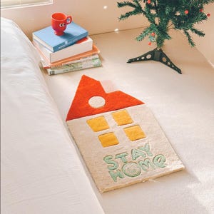 May include: A white rug with a red and yellow house design and the words "STAY HOME" in green. The rug is on a white bed with a stack of books and a red cup on top.