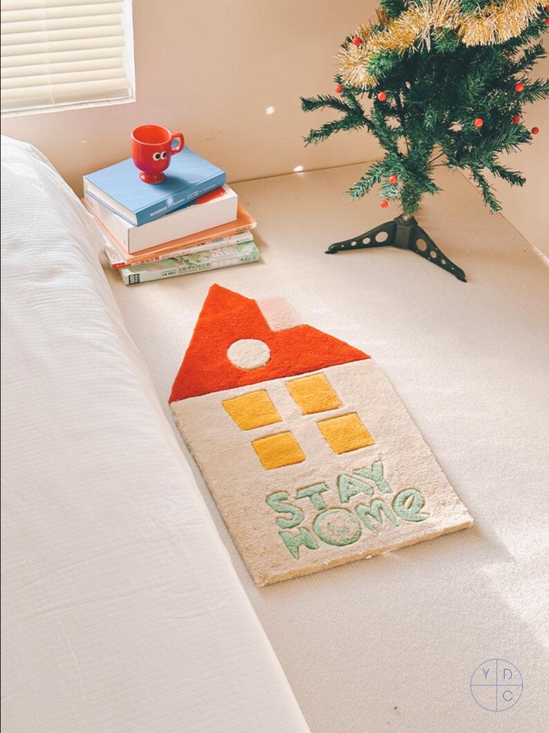 May include: A white rug with a red and yellow house design and the words "STAY HOME" in green. The rug is on a white bed with a stack of books and a red cup on top.