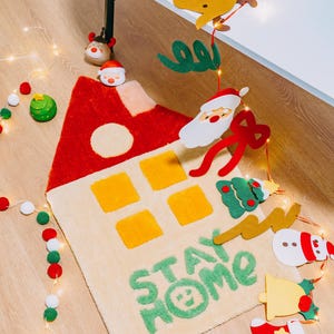 May include: A beige rug with a red and yellow house design and the words "Stay Home" in green. The rug is surrounded by Christmas decorations, including a snowman, a Santa Claus, a reindeer, and a Christmas tree.