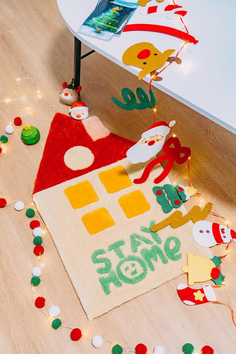 May include: A beige rug with a red and yellow house design and the words "Stay Home" in green. The rug is surrounded by Christmas decorations, including a snowman, a Santa Claus, a reindeer, and a Christmas tree.