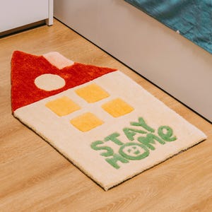 May include: A cream-colored rug shaped like a house with a red roof, yellow windows, and the words "STAY HOME" in green.