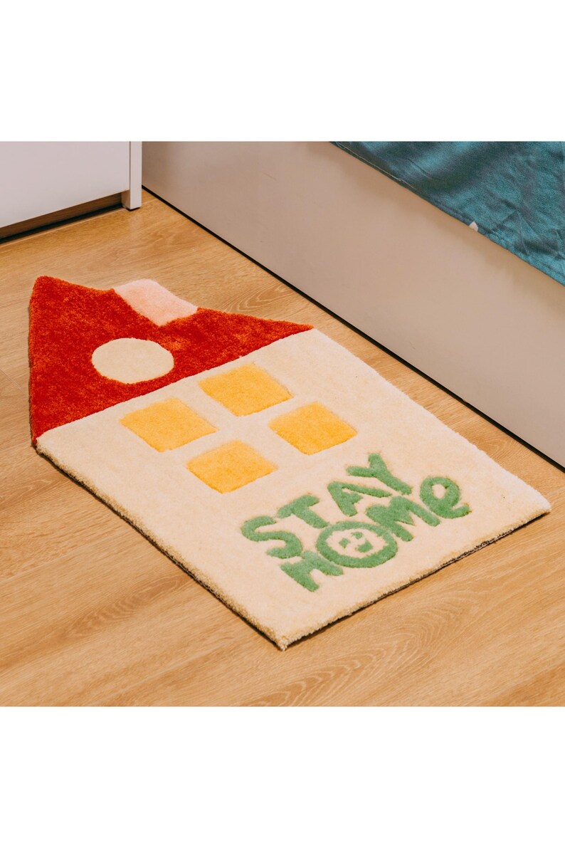 May include: A cream-colored rug shaped like a house with a red roof, yellow windows, and the words "STAY HOME" in green.
