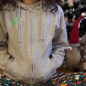 Front view of grey zip hoodie with small green chakra symbol on chest, relaxed cozy home look