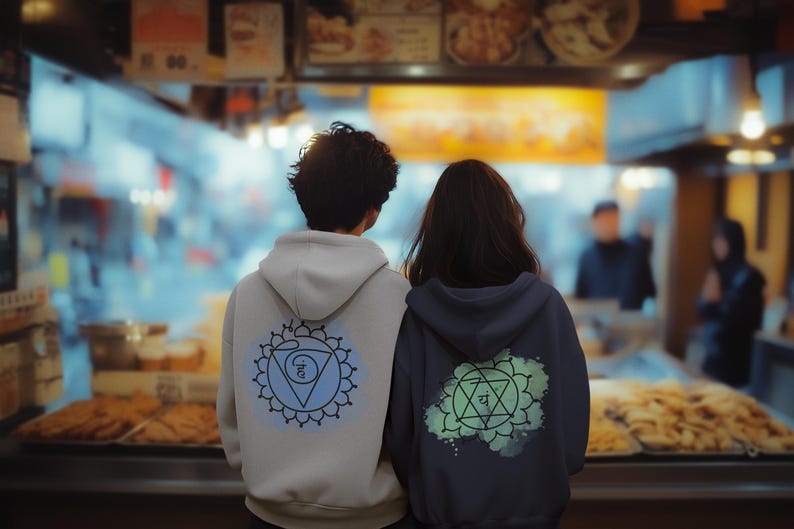 A couple explores a vibrant Asian market, wearing hoodies with large Anahata chakra prints on the back. The man wears black, and the woman wears gray. The scene symbolizes love, balance, and conneсtion