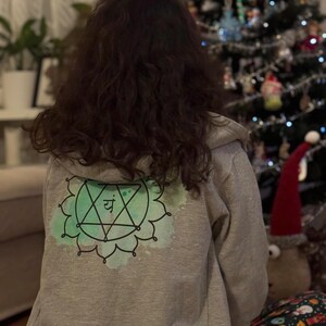 Back view of grey spiritual hoodie featuring green heart chakra symbol, holiday living room background