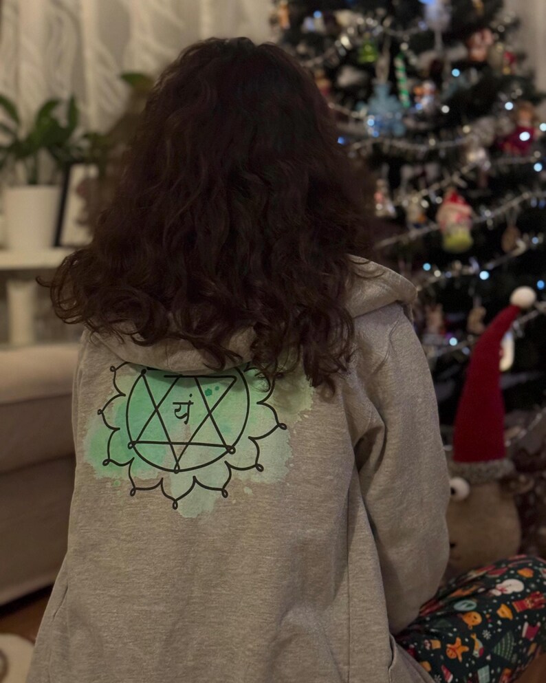 Back view of grey spiritual hoodie featuring green heart chakra symbol, holiday living room background