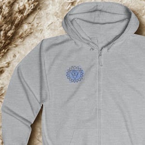 Light gray unisex zip hoodie featuring blue chakra mandala on the chest. Minimal spiritual hoodie presented in flat lay composition, designed for mindful everyday wear and inner balance.