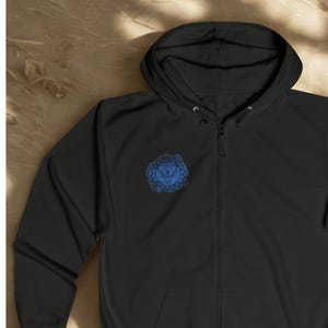 Black unisex zip hoodie shown in flat lay style with blue chakra mandala printed on the chest. Minimal spiritual zip up hoodie with calm energy design, photographed on natural textured background.