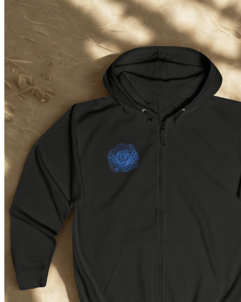 Black unisex zip hoodie shown in flat lay style with blue chakra mandala printed on the chest. Minimal spiritual zip up hoodie with calm energy design, photographed on natural textured background.