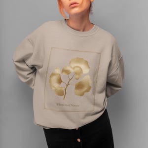 May include: A beige sweatshirt featuring a gold-colored ginkgo leaf design within a square frame. The design includes a branch with multiple leaves. The text "Whispers of Nature" is below the design. The sweatshirt is a neutral color.