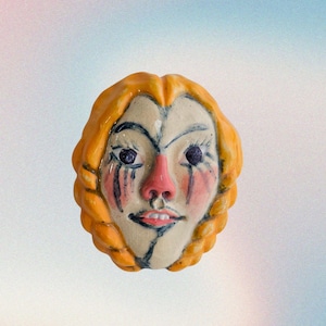 May include: A ceramic face sculpture with orange hair and a painted face. The face has dark blue eyes, a pink nose, and red tear streaks. The sculpture has a slightly open mouth and is set against a soft, gradient background.