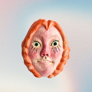 May include: A whimsical, hand-painted ceramic face with orange hair and rosy cheeks. The face has large green eyes, a small nose, and a slight smile. The background is a soft gradient of blue and pink.
