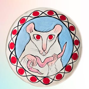 May include: A white ceramic plate with a whimsical illustration of a rat holding its tail. The rat has red eyes and pink ears and tail. The background is light blue, and the plate is bordered with red circles.