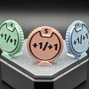 Könnte beinhalten: Drei mehrfarbige Dreh-Zähler mit den Zahlen 4, 5 und 19. Jeder Zähler hat ein +1/+1-Symbol. Die Zähler sind grün, pfirsichfarben und blau. Die Zähler befinden sich auf einer grauen Oberfläche.
