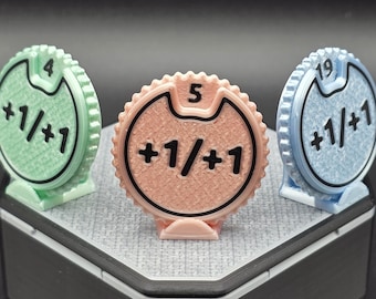 May include: Three multi-colored spin counters with the numbers 4, 5, and 19. Each counter has a +1/+1 symbol. The counters are in green, peach, and blue. The counters are on a gray surface.