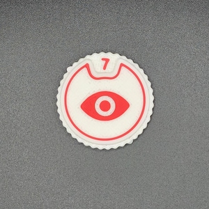 May include: A white and red circular emblem with a scalloped edge. The emblem features a red eye symbol and the number 7 in a red font. The background is a textured grey surface.