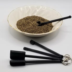Peut inclure: Quatre mini pelles noires avec un porte-clés argenté attaché à l'une des poignées. Les pelles sont dans un bol rempli de grains de poivre brun.