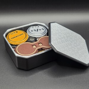 May include: A black, rectangular box with a silver lid, containing game tokens. The tokens include a yellow coin with "TREASURE" printed on it, a silver coin with "+1/+1", and a brown token with the number "4" on it.
