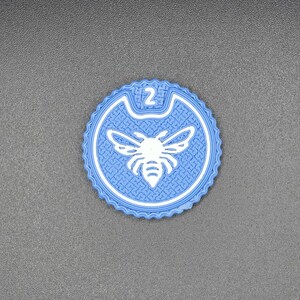 Peut inclure: Un dessous de verre rond bleu clair avec un motif d'abeille blanc au centre. Le dessous de verre a une surface texturée et une bordure blanche. Le chiffre "2" est en haut.